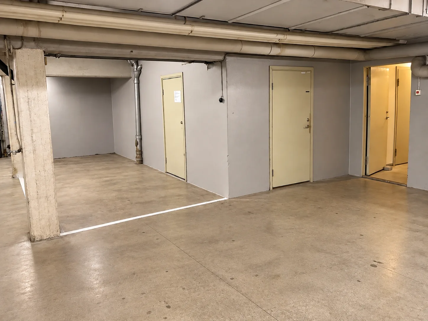 Källarutrymme med ljusgrå väggar, beige metalldörrar, betongpelare och rör i taket