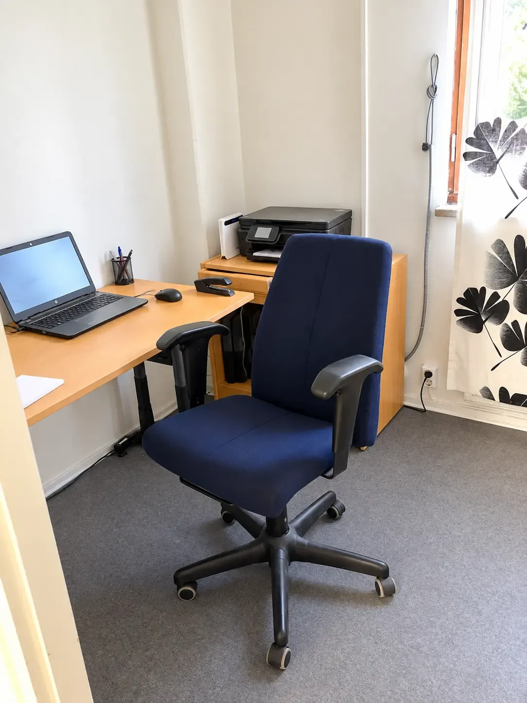 Kontorsrum med skrivbord, bärbar dator, kontorsstol och skrivare vid fönster