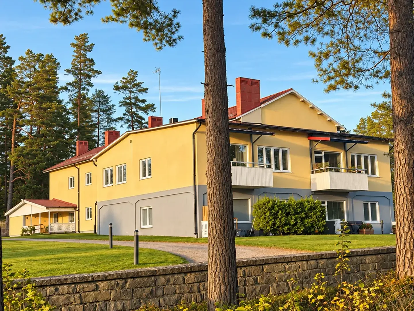 Norra Storgatan 10A–B i sen eftermiddagssol sedd från grusvägen med gräsmatta, låg stenmur och höga tallar i förgrunden