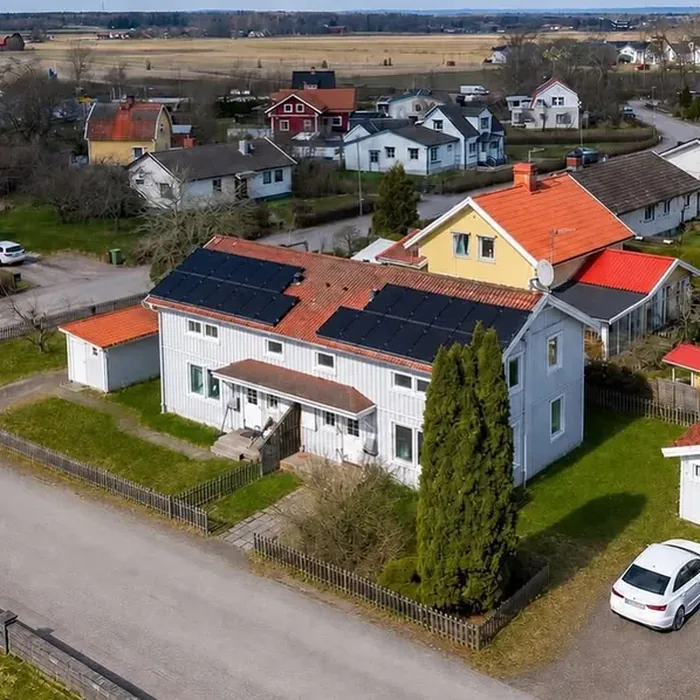 Flygfoto av en ny fastighet: ett ljusgrått tvåvåningshus med tegelrött sadeltak och solceller, omgivet av trädgård och öppna fält.